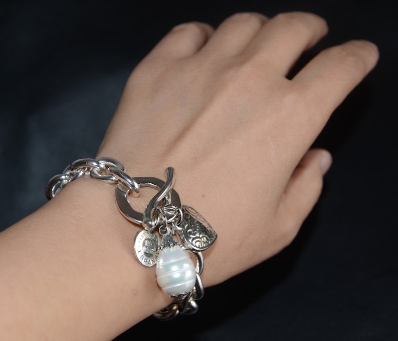 Puede incluir: Un brazalete de plata con un gran encanto de perla, un encanto en forma de coraz&oacute;n y otros encantos m&aacute;s peque&ntilde;os. El brazalete se lleva en la mu&ntilde;eca.
