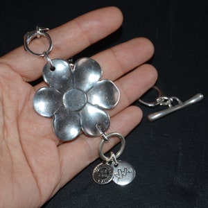 以下が含まれることがあります： 大きな花のチャーム、小さな「Made with Love」の文字が刻まれたチャーム、そしてトグル留め具が付いたシルバーブレスレットです。