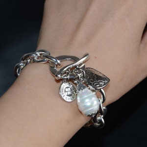 Puede incluir: Un brazalete de plata con un gran eslab&oacute;n de cadena de plata, un dije de coraz&oacute;n, un dije de perla y un dije con el texto "Hecho en Espa&ntilde;a".