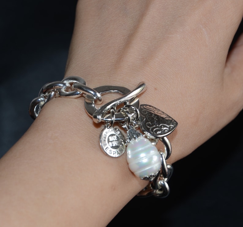 Puede incluir: Un brazalete de plata con un gran eslab&oacute;n de cadena de plata, un dije de coraz&oacute;n, un dije de perla y un dije con el texto "Hecho en Espa&ntilde;a".