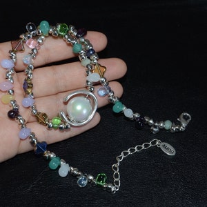 Op de afbeelding: Een veelkleurige kralenketting met een grote witte parel hanger. De ketting heeft een zilveren ketting met een klein label waarop "AMOR" staat.