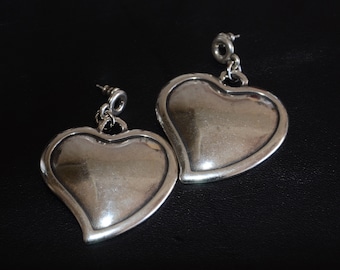 May include: A pair of silver heart-shaped dangle earrings. The earrings have a smooth, polished finish and are suspended from a small loop.