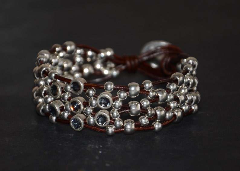 May include: A brown leather bracelet with multiple strands of silver beads and dark blue crystal accents.