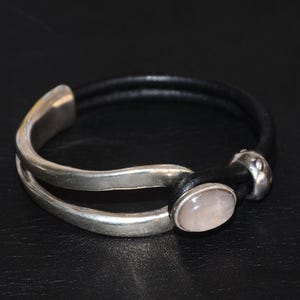 May include: A silver and black bracelet with a rose quartz stone. The bracelet features a double-strand black leather band and a silver clasp. The rose quartz stone is oval-shaped and set in a silver bezel.