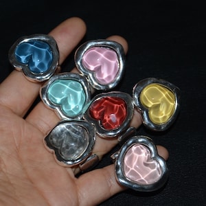 May include: Seven rings with heart-shaped, colourful, translucent centres. Each ring has a silver-coloured, thick band and bezel. The heart-shaped centres are in shades of blue, pink, green, red, yellow, and clear.