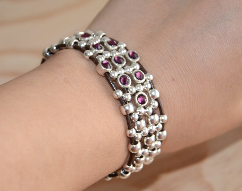 May include: A multi-strand bracelet with brown leather cord and silver beads accented with purple gemstones.