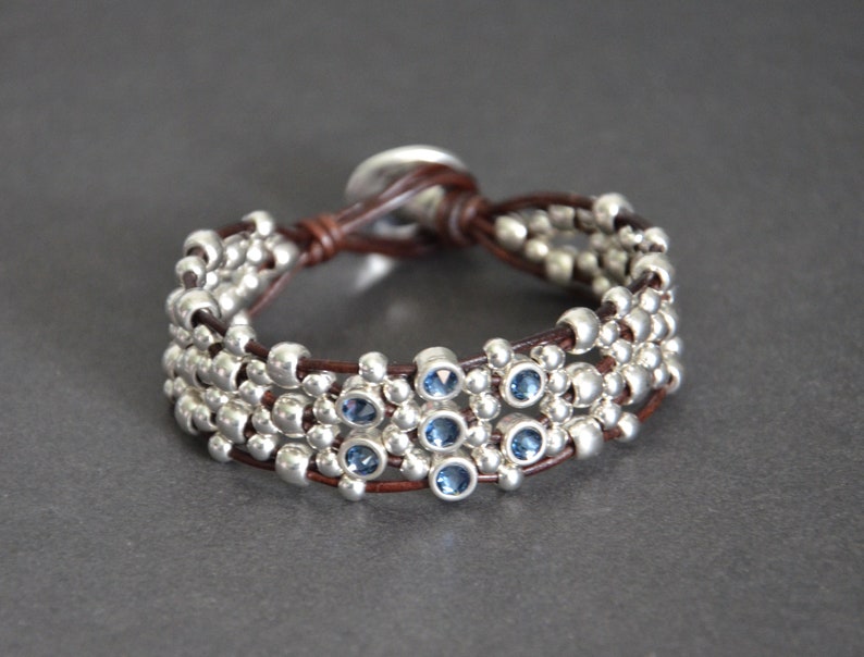 May include: A brown leather bracelet with silver beads and blue gemstones. The bracelet is adjustable with a sliding knot.