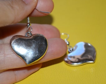 May include: A pair of silver heart-shaped earrings. Each earring features a polished, rounded heart pendant suspended from a silver fish hook. The earrings are displayed against a bright yellow background.