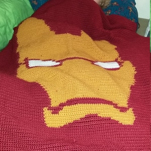 Può includere: Una coperta all'uncinetto rossa con un disegno di viso di Iron Man giallo. Un bambino è parzialmente nascosto sotto la coperta.