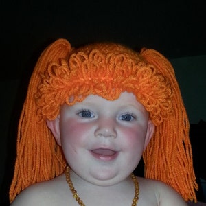 Könnte beinhalten: Ein Kind trägt eine leuchtend orangefarbene gehäkelte Perücke mit zwei langen Zöpfen. Die Perücke ist aus Garn gefertigt und hat eine lockige Textur. Das Kind trägt auch eine goldene Halskette.