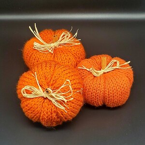 以下が含まれることがあります： 茶色の茎とラフィアのリボンが付いた、オレンジ色のニットの南瓜が3つ。南瓜は黒い表面に並べられています。
