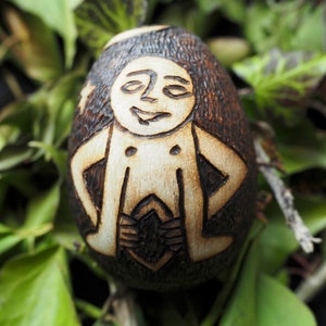 May include: A wooden egg with a carved design of a person with a smiling face. The egg is brown and the carving is in a light brown color.