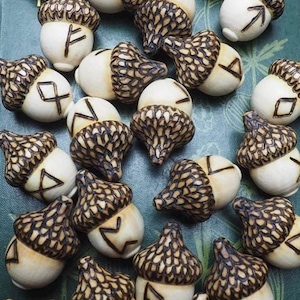 May include: A collection of 25 acorn-shaped charms with different runes carved into them. The acorns are made of wood and have a light brown color. They are arranged on a green book with a black and white patterned background.