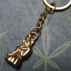 May include: A gold-colored keychain featuring a small, detailed figurine of a person. The keychain is attached to a gold-colored metal ring.