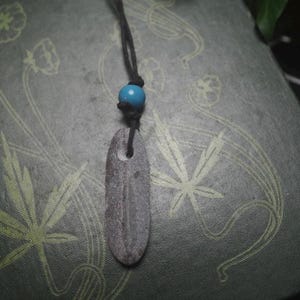 May include: A pendant necklace featuring a smooth, elongated gray stone with a natural hole near the top, suspended from a black cord. A single, round, bright blue bead is threaded onto the cord just above the stone, securing the pendant in place.