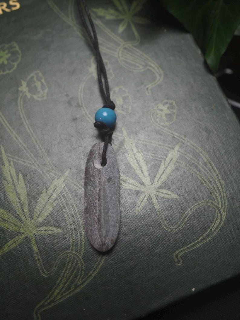 May include: A pendant necklace featuring a smooth, elongated gray stone with a natural hole near the top, suspended from a black cord. A single, round, bright blue bead is threaded onto the cord just above the stone, securing the pendant in place.