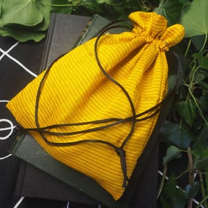 May include: A bright yellow drawstring pouch featuring thin, dark vertical pinstripes. The bag is cinched at the top with two long, thin black cords that hang down, creating a simple closure mechanism for the fabric container.