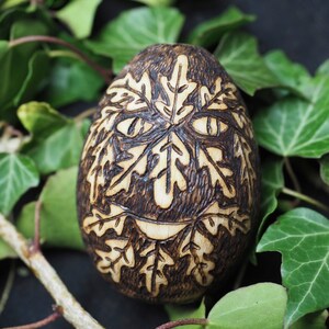 May include: A wooden egg with a carved face and leaf design. The egg is dark brown and has a light brown face with a smile. The egg is surrounded by green leaves.