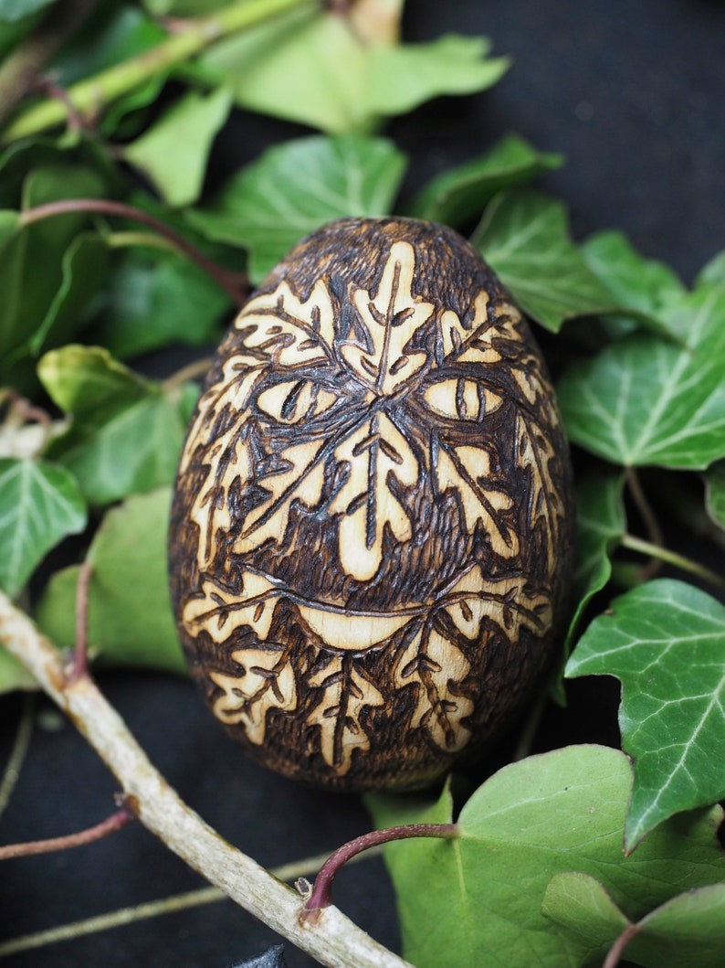 May include: A wooden egg with a carved face and leaf design. The egg is dark brown and has a light brown face with a smile. The egg is surrounded by green leaves.
