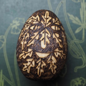 May include: A wooden egg with a woodburned design of a face with leaves. The face is smiling and has a whimsical look.