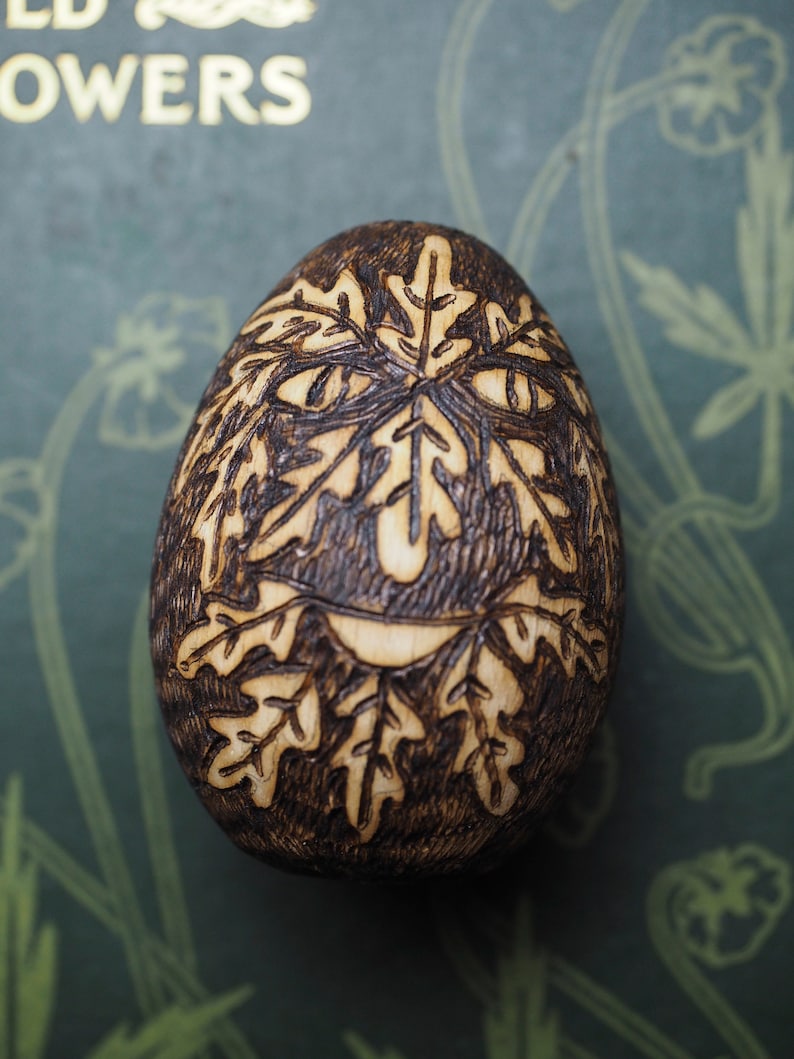May include: A wooden egg with a woodburned design of a face with leaves. The face is smiling and has a whimsical look.