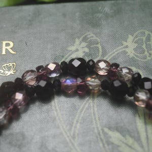 May include: A multi-strand beaded bracelet featuring faceted black beads interspersed with translucent purple and iridescent clear crystal beads. The beads are arranged in a repeating pattern, creating a textured appearance with light reflecting off the surfaces.