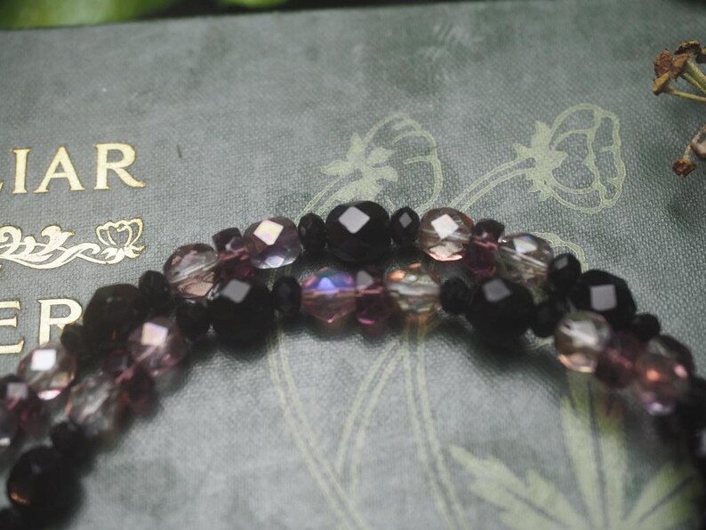 May include: A multi-strand beaded bracelet featuring faceted black beads interspersed with translucent purple and iridescent clear crystal beads. The beads are arranged in a repeating pattern, creating a textured appearance with light reflecting off the surfaces.