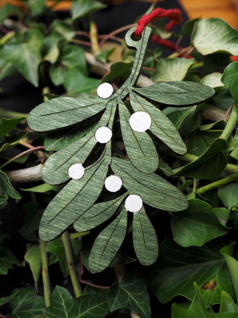 May include: A green wooden mistletoe sprig with white painted berries. The sprig is hanging from a red string.
