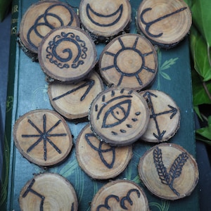 May include: A collection of wooden rune stones with various black symbols, including a crescent moon, sun, and eye. The circular stones are arranged on a green book, with green leaves in the background. The runes are made of natural wood.