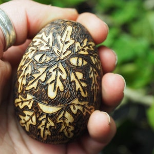 May include: A wooden egg with a carved design of leaves and a face. The egg is brown and has a light brown wood grain. The face is made up of leaves and has two eyes and a mouth.