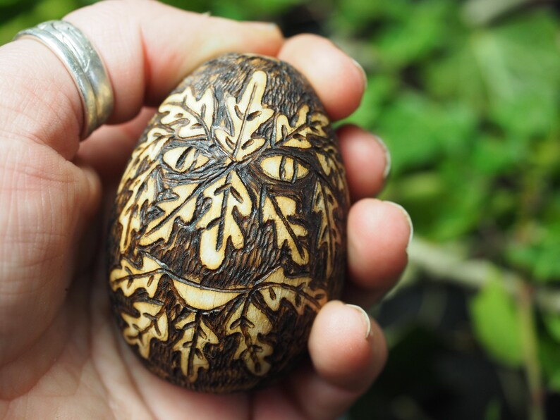 May include: A wooden egg with a carved design of leaves and a face. The egg is brown and has a light brown wood grain. The face is made up of leaves and has two eyes and a mouth.