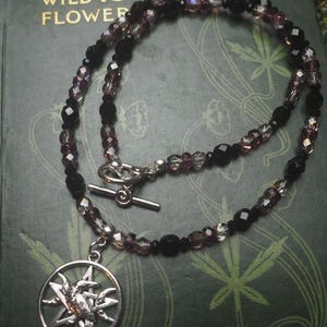 May include: A beaded necklace featuring alternating black and translucent purple faceted beads with a silver-toned toggle clasp. A circular silver-toned pendant hangs from the necklace, depicting a bird silhouette centered within a five-pointed star.