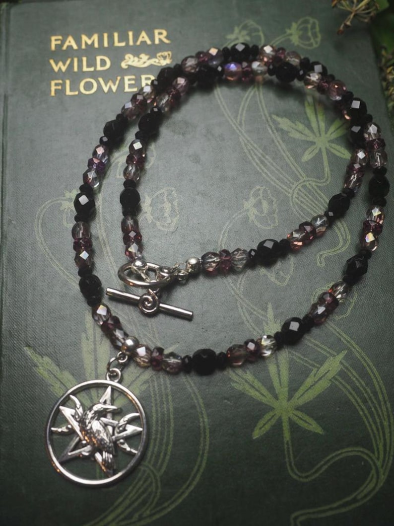 May include: A beaded necklace featuring alternating black and translucent purple faceted beads with a silver-toned toggle clasp. A circular silver-toned pendant hangs from the necklace, depicting a bird silhouette centered within a five-pointed star.