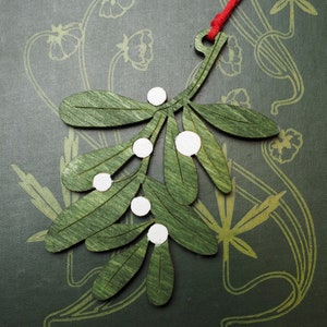 May include: A green wooden mistletoe sprig with white painted berries hanging from a red string. The mistletoe is a traditional Christmas decoration.