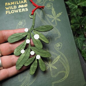 May include: A green wooden mistletoe sprig ornament with white painted berries. The ornament is hanging from a red string.
