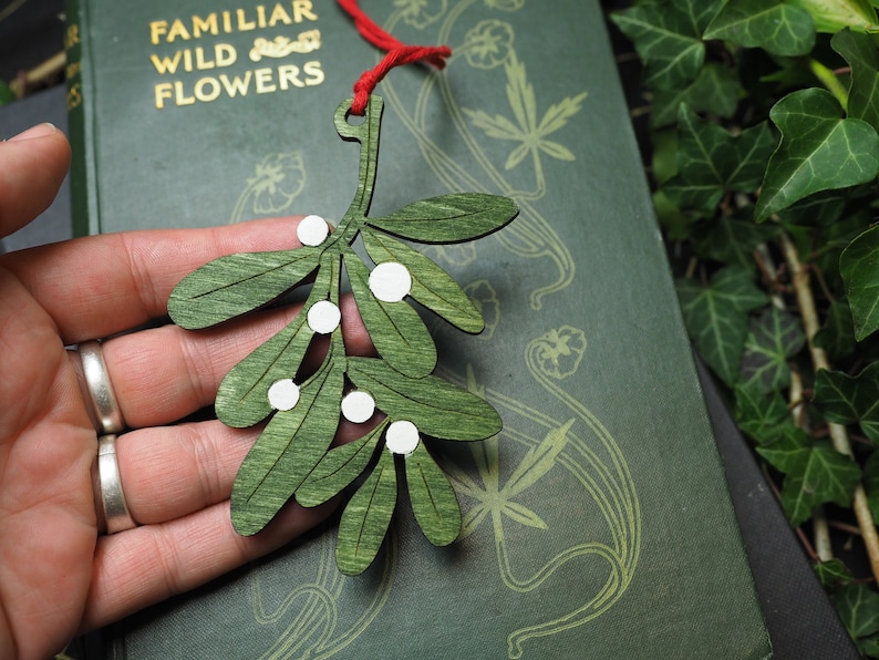 May include: A green wooden mistletoe sprig ornament with white painted berries. The ornament is hanging from a red string.