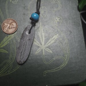 May include: A dark gray, elongated, flat stone pendant with a hole at the top, suspended from a black cord featuring a single bright blue bead. A copper-colored United States penny is positioned to the left for scale, showing the relative size.