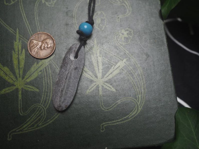 May include: A dark gray, elongated, flat stone pendant with a hole at the top, suspended from a black cord featuring a single bright blue bead. A copper-colored United States penny is positioned to the left for scale, showing the relative size.