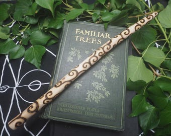 Può includere: Una bacchetta in legno chiaro caratterizzata da un design a pirografia marrone scuro con spirali e volute ripetute. La bacchetta è posizionata diagonalmente sopra un libro con copertina rigida verde scuro intitolato Familiar Trees with Coloured Plates.