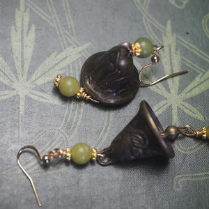 May include: A pair of antique-style earrings featuring bell-shaped pendants with intricate designs. Each earring has a gold-colored hook and is adorned with a light green bead and gold-colored accents. The earrings rest on a green surface.