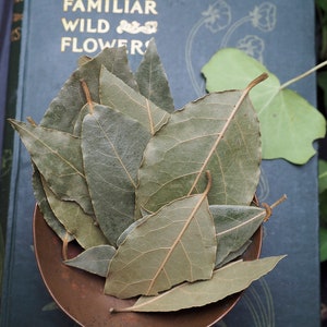 May include: A pile of dried bay leaves in a copper bowl. The leaves are green and brown and have a distinct veined pattern. The bowl is sitting on a book with the title "Familiar Wild Flowers" visible.