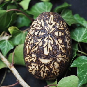 May include: A wooden egg with a carved design of leaves and a face. The egg is brown and has a natural wood finish. It is surrounded by green leaves.