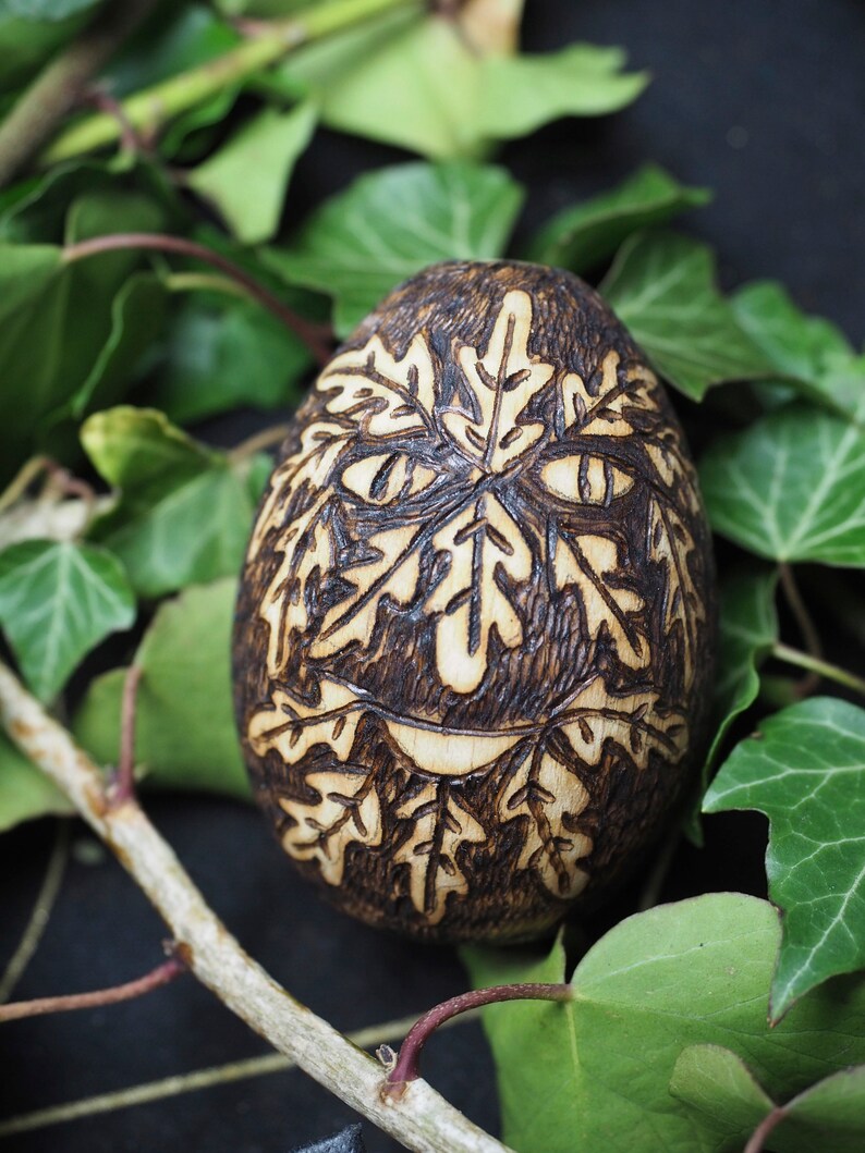 May include: A wooden egg with a carved design of leaves and a face. The egg is brown and has a natural wood finish. It is surrounded by green leaves.
