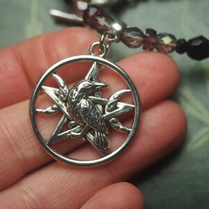 May include: A silver-toned circular pendant featuring a five-pointed star with a raven perched in the center and a crescent moon on the side. The pendant is attached to a beaded bracelet strand made of dark purple and black faceted beads.