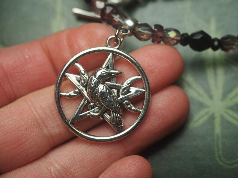 May include: A silver-toned circular pendant featuring a five-pointed star with a raven perched in the center and a crescent moon on the side. The pendant is attached to a beaded bracelet strand made of dark purple and black faceted beads.