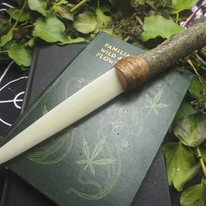 May include: A handcrafted knife with a light-colored blade and a handle made from a tree branch. The handle is wrapped with twine. The knife rests on a dark green book with the title "FAMILIA WILD & FLOW". Green leaves surround the knife and book.