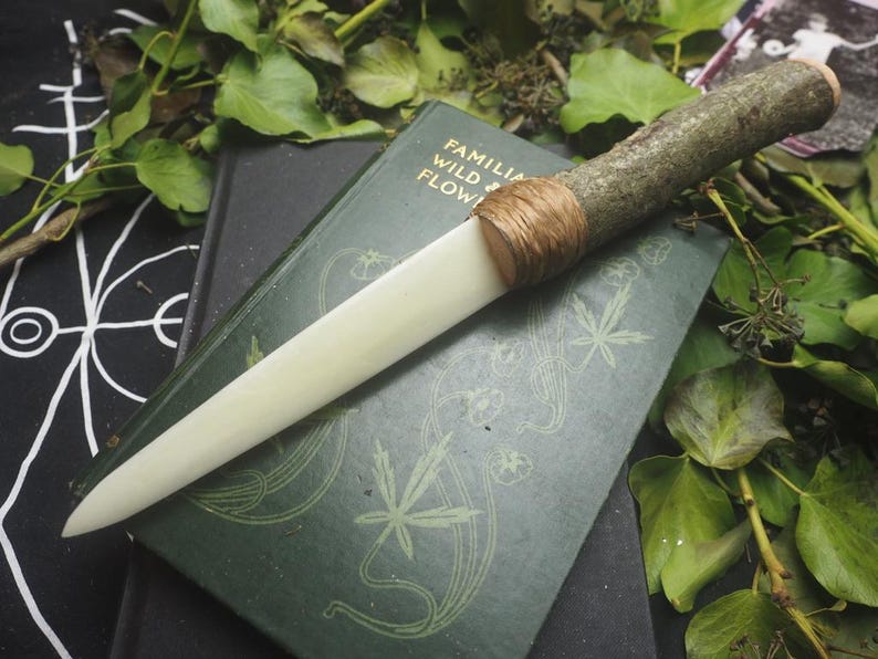May include: A handcrafted knife with a light-colored blade and a handle made from a tree branch. The handle is wrapped with twine. The knife rests on a dark green book with the title "FAMILIA WILD & FLOW". Green leaves surround the knife and book.