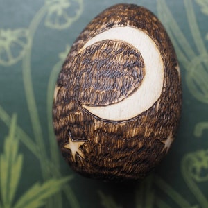 May include: A wooden egg with a crescent moon and stars carved into it. The egg is dark brown and the carving is light brown.