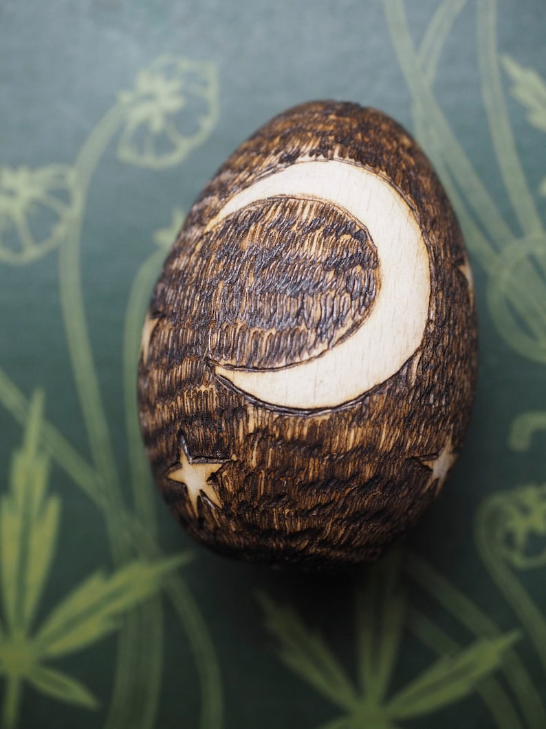 May include: A wooden egg with a crescent moon and stars carved into it. The egg is dark brown and the carving is light brown.
