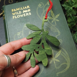 May include: A green wooden mistletoe sprig ornament with a red string attached. The ornament is held in a hand against a green book with the text "Familiar Wild Flowers" on the cover.
